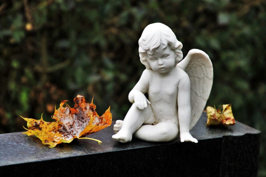 mały porcelanowy biały aniołek na granitowym pomniku. obok leżą pomarańczowe jesienne liście