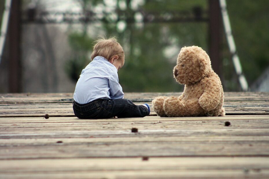 małe dziecko siedzi na drewnianym pomoście. na przeciw niego siedzi duży pluszowy miś.