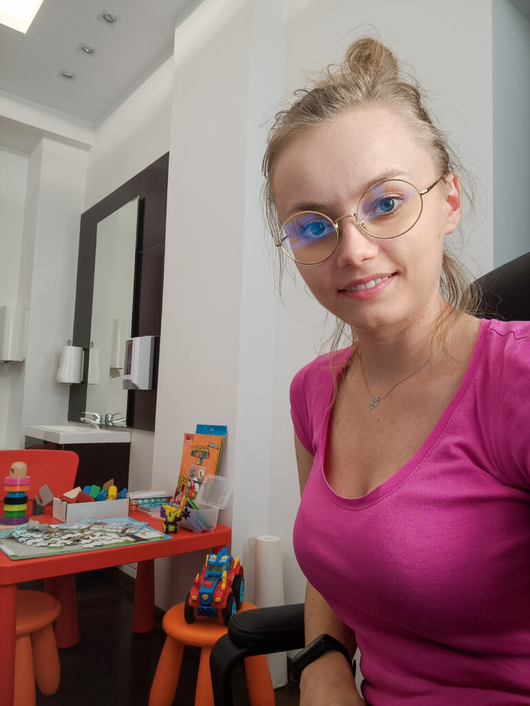 Julia Skibińska w różowej bluzce i związanych w koczek włosach. w tle gabinet ze stolikiem dziecięcym i zabawkami