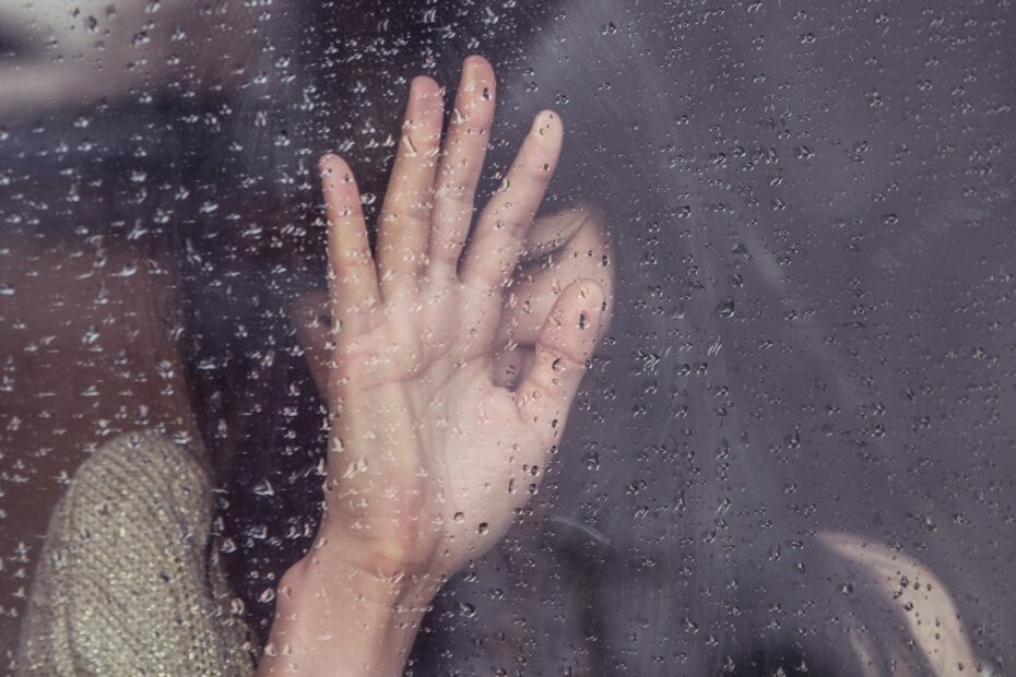 czarnowłosa kobieta stoi za szybą. pada deszcz. krople deszczu osadzają się na szybie. kobieta trzyma dłoń na na szybie wnętrzem do niej zasłaniając swoją twarz