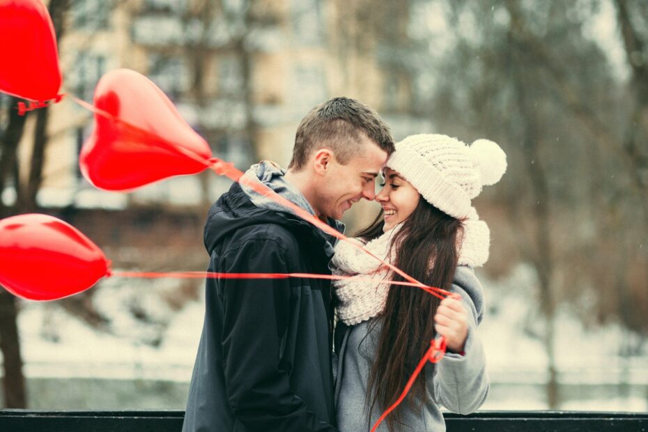 młody mężczyzna i młoda kobieta ubrani w kurtki uśmiechają się patrząc na siebie i stykając nosami. kobieta trzyma w lewej dłoni 3 czerwone balony w kształcie serc.