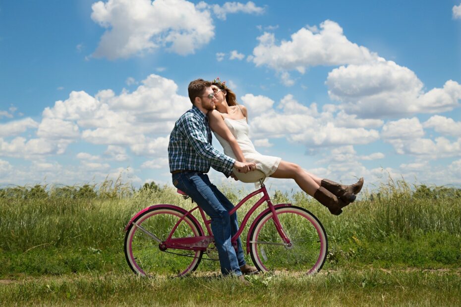 Para na tle zielonej trawy i błękitnego nieba z chmurami. mężczyzna w jeansach i niebieskiej koszuli w kratę siedzi na ciemnoróżowym rowerze. dziewczyna w białej sukience i brązowych butach siedzi na kierownicy tego roweru i daje buziaka chłopakowi
